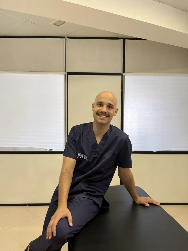 Eduard Folch, fisioterapeuta, sonriente y sentado en una camilla de la consulta con el uniforme de Fisio Folch